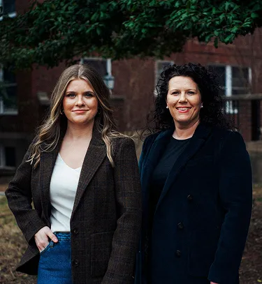 Two women standing outdoors in front of a brick building, wearing dark blazers and smiling.