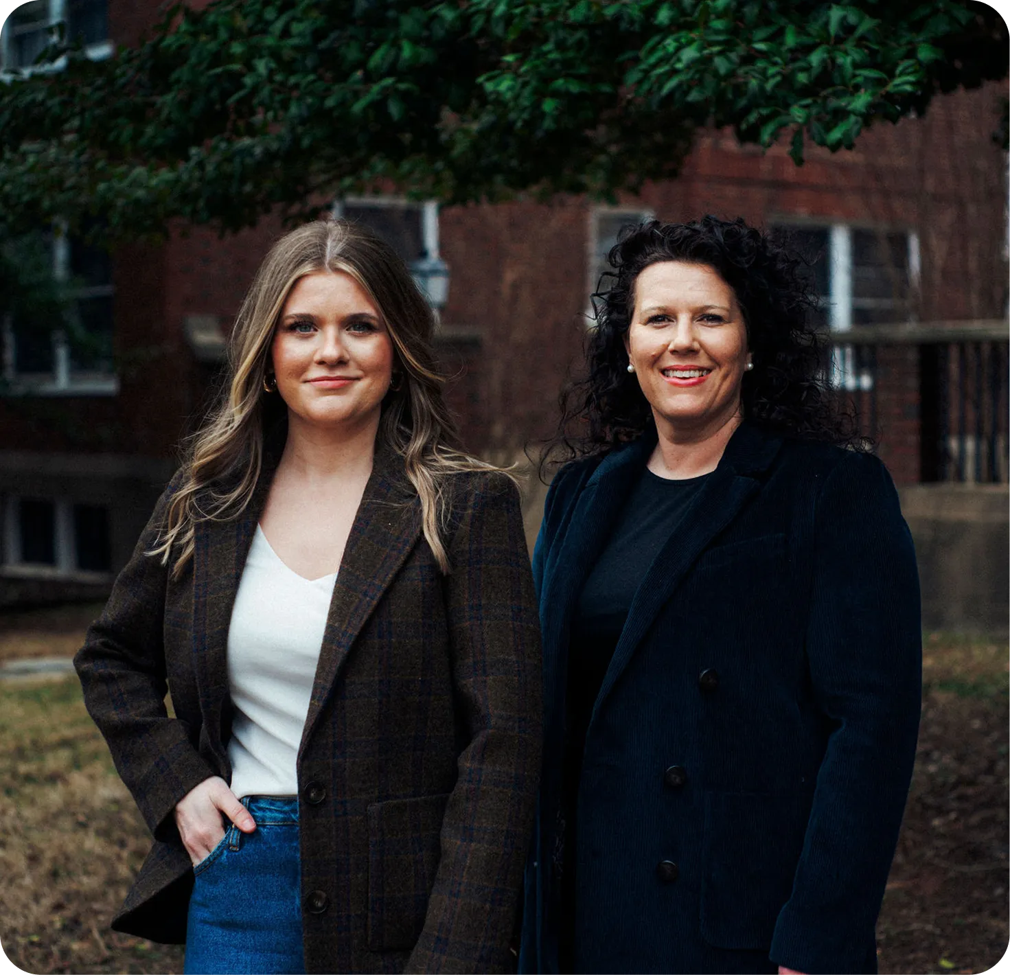 Two professional women standing outdoors, one with blonde hair in a brown plaid blazer and jeans, the other with curly dark hair in a black coat, with a brick building and greenery in the background.