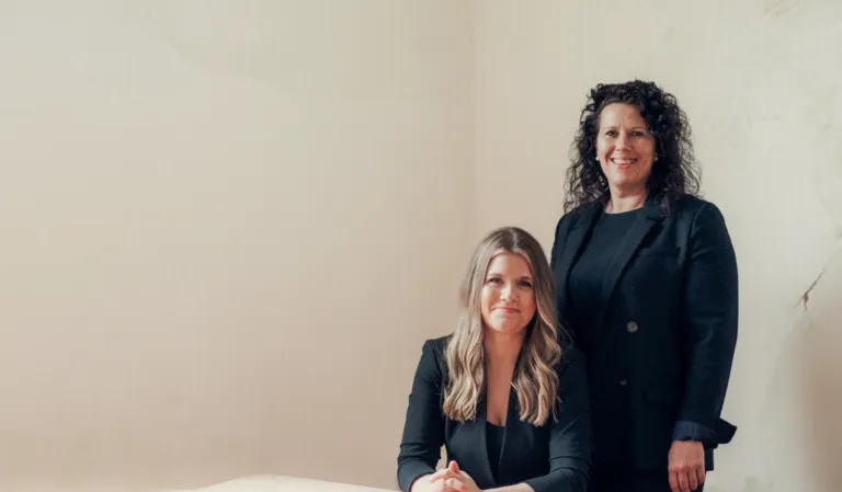 Two professional women in black blazers posing against a plain beige wall.