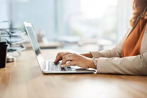 Person typing on a silver laptop at a wooden desk in a bright office setting.