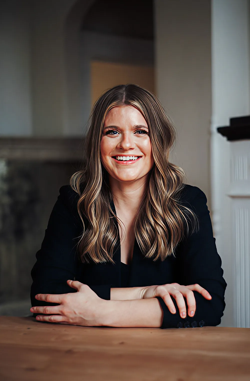 Portrait of a smiling woman with long wavy hair, wearing a black blazer, seated at a wooden table.