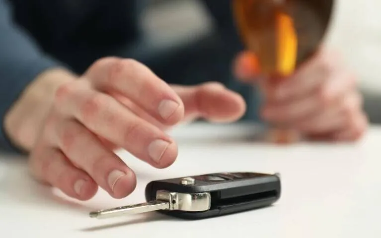 Close-up of a hand reaching for a car key on a white table, holding a glass of amber liquid.