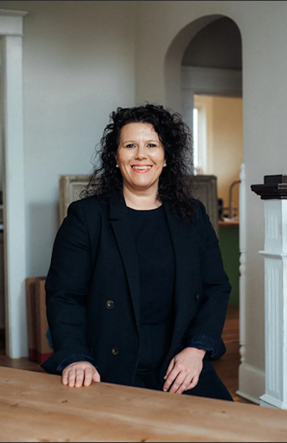 Professional woman with curly dark hair wearing a black blazer, seated indoors smiling.