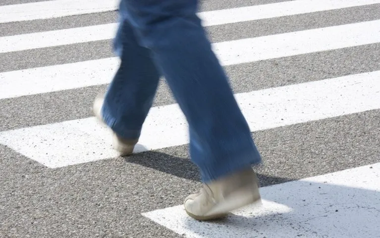 Person wearing blue jeans and beige shoes walking across a white-striped pedestrian crosswalk on asphalt.