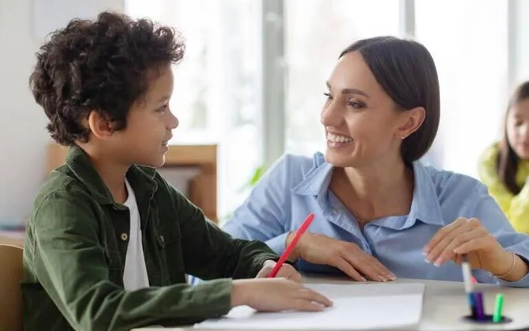 Teacher engaging with a young student at a desk, both smiling and holding pens.