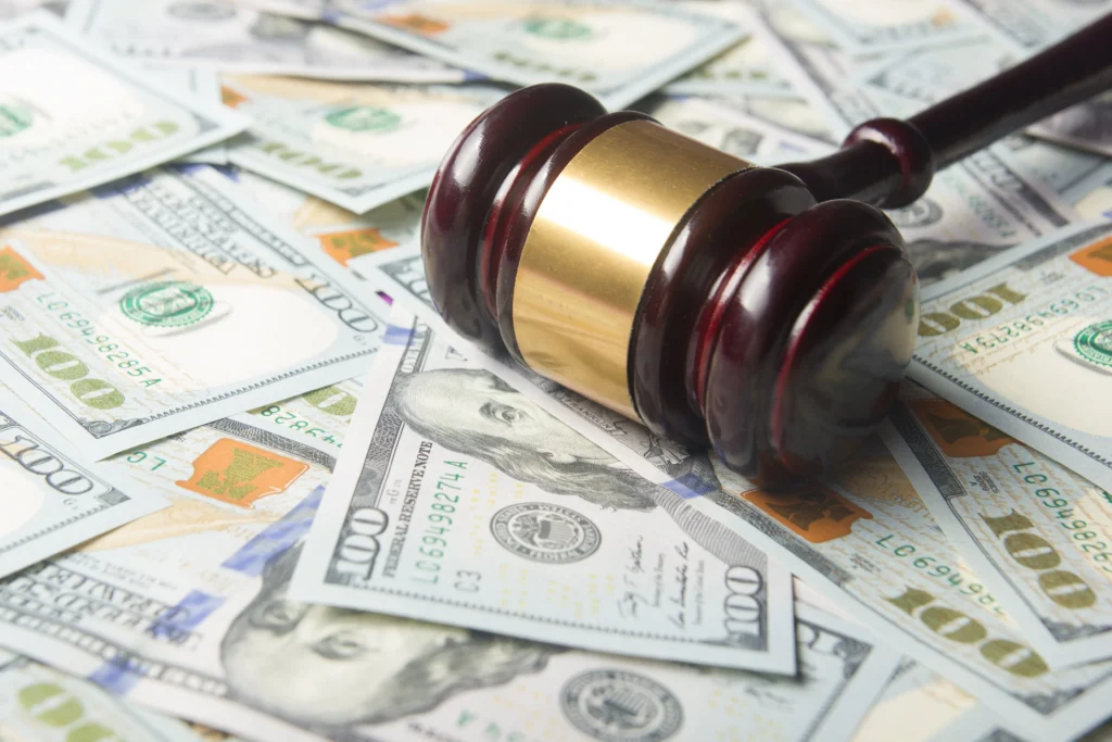 Wooden judge's gavel with gold band resting on scattered U.S. hundred-dollar bills.