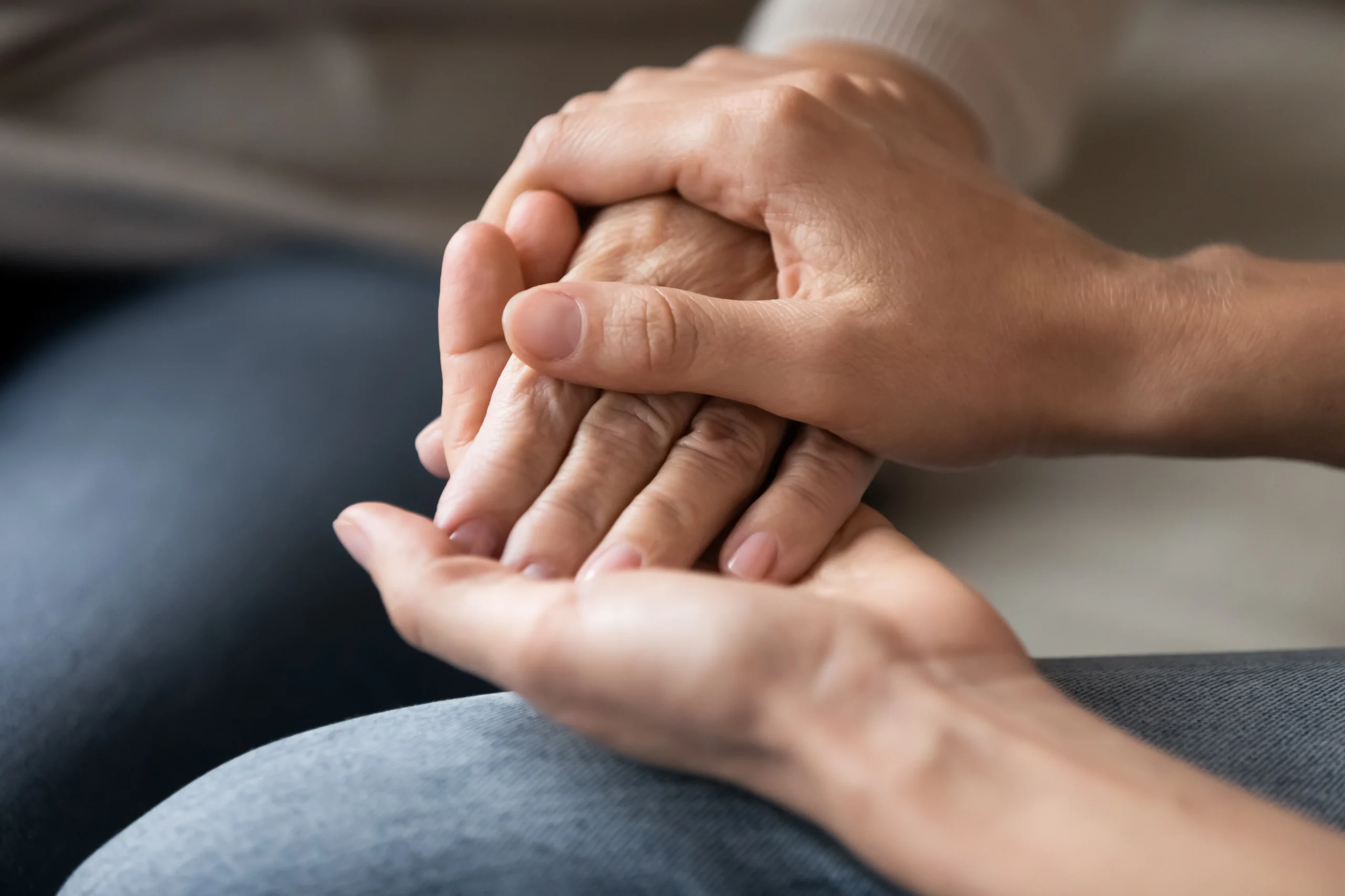 Close-up of a younger hand gently holding and supporting an elderly hand over a gray fabric surface.