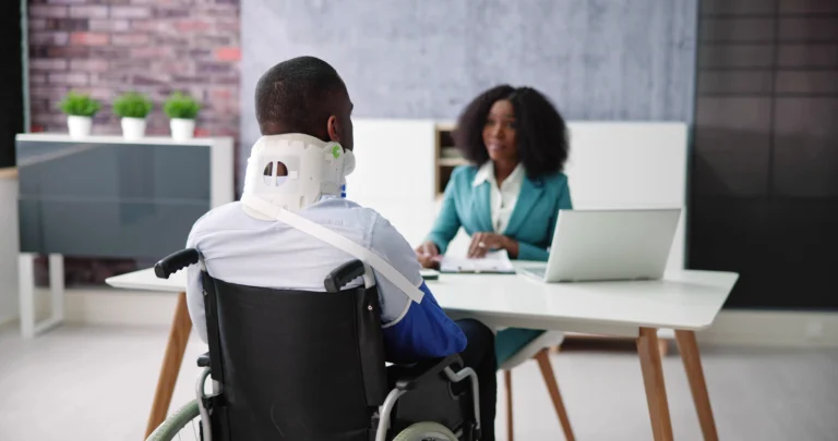 Man in wheelchair wearing neck brace consulting female professional in teal blazer at office desk.