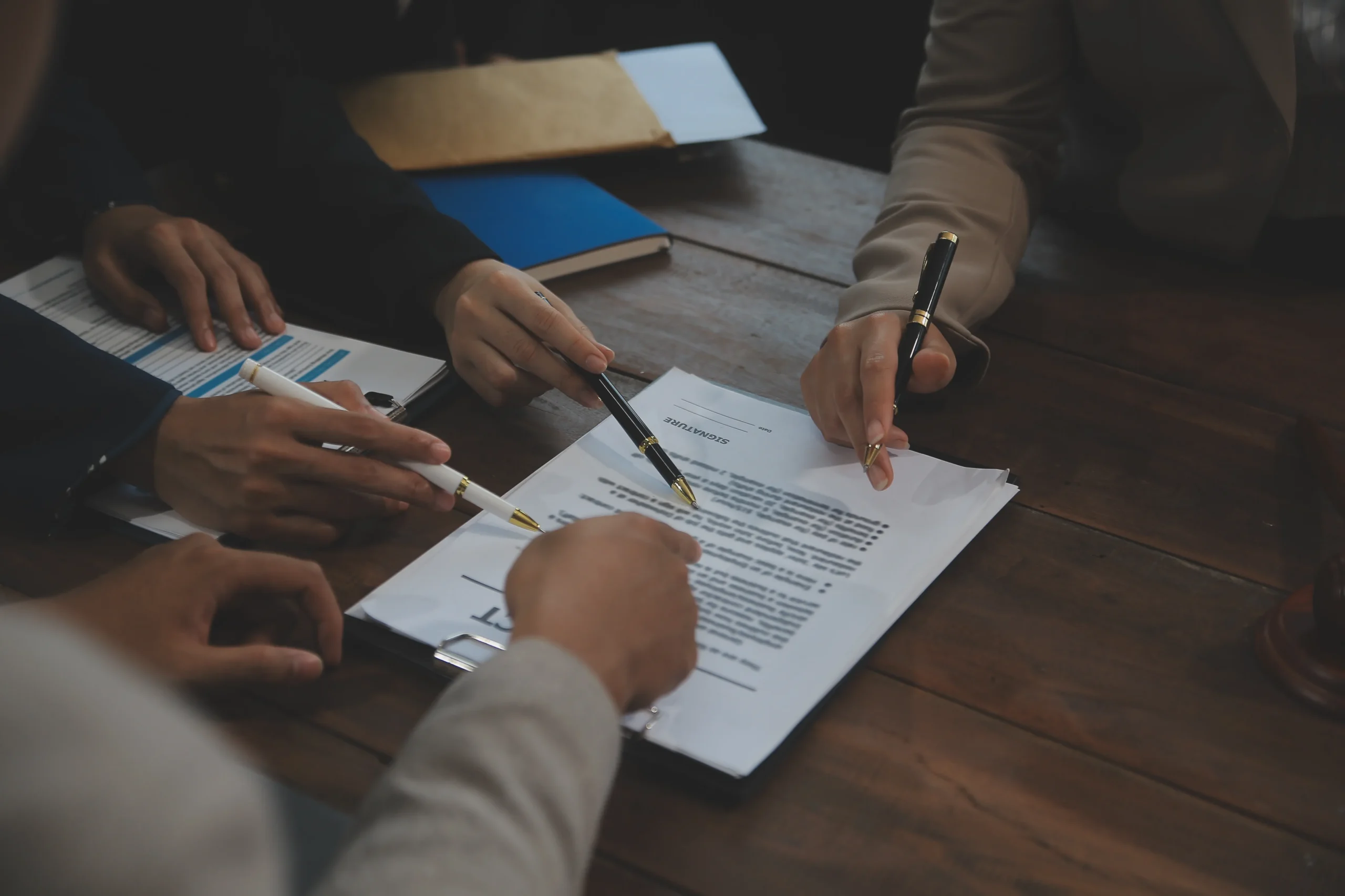 Close-up of three people reviewing and pointing at contract documents on a wooden table.