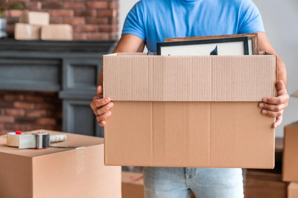 Person in blue shirt holding cardboard box with picture frames inside, surrounded by moving supplies.
