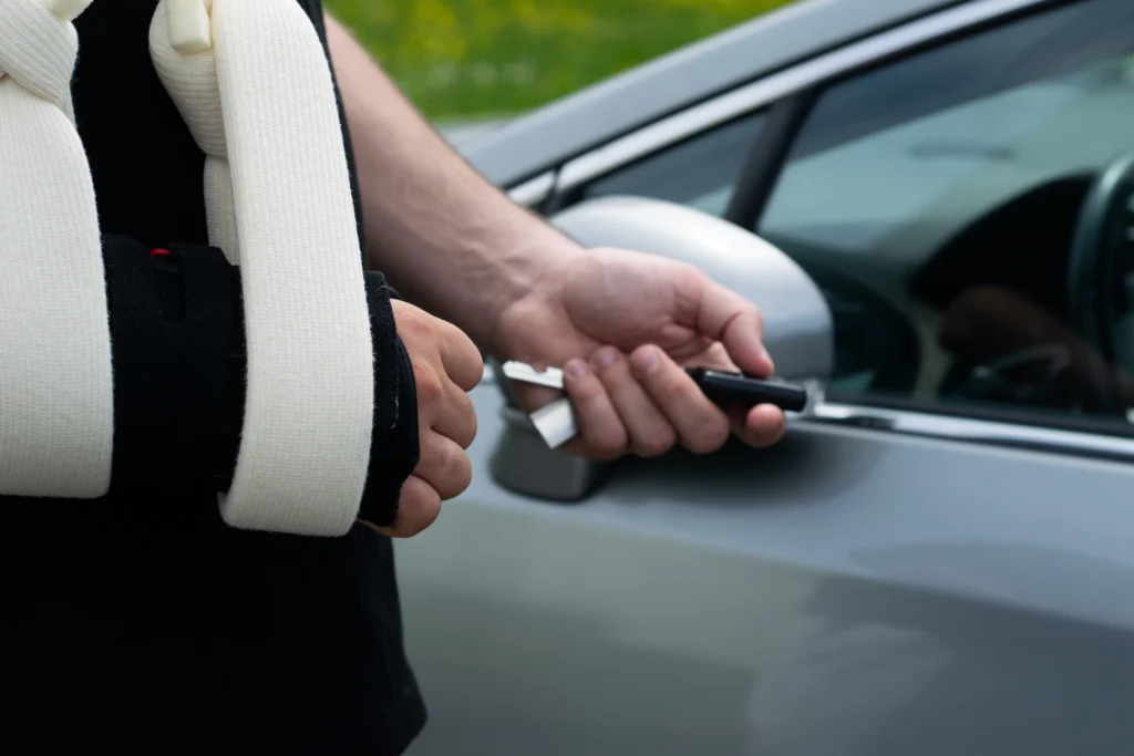 Person with arm in white sling holding car key near silver vehicle side mirror outdoors.