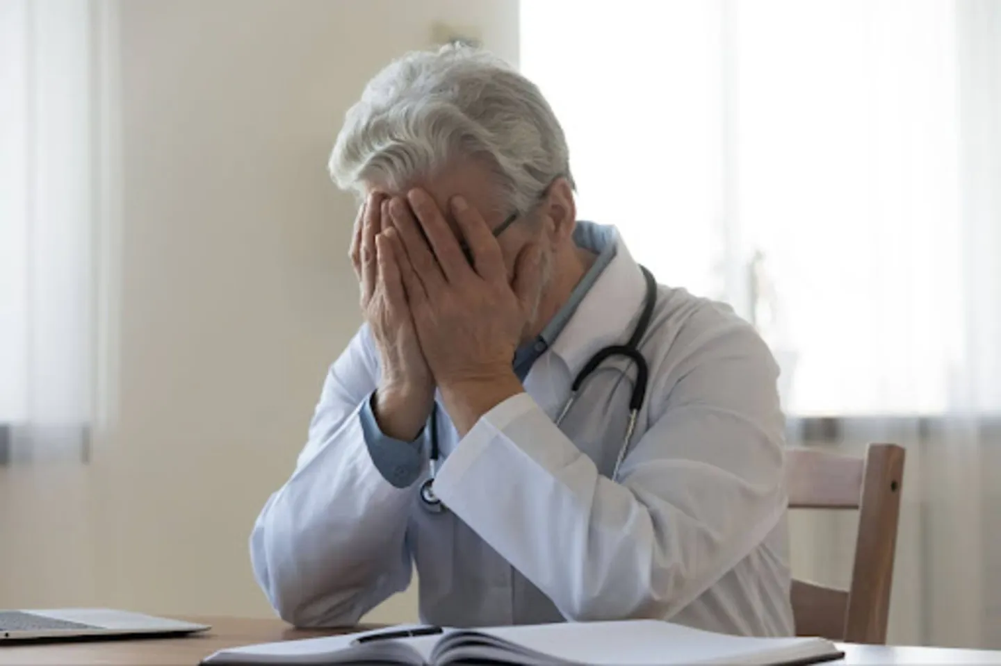 Elderly male doctor in white coat sitting at desk, covering face with hands in distress.