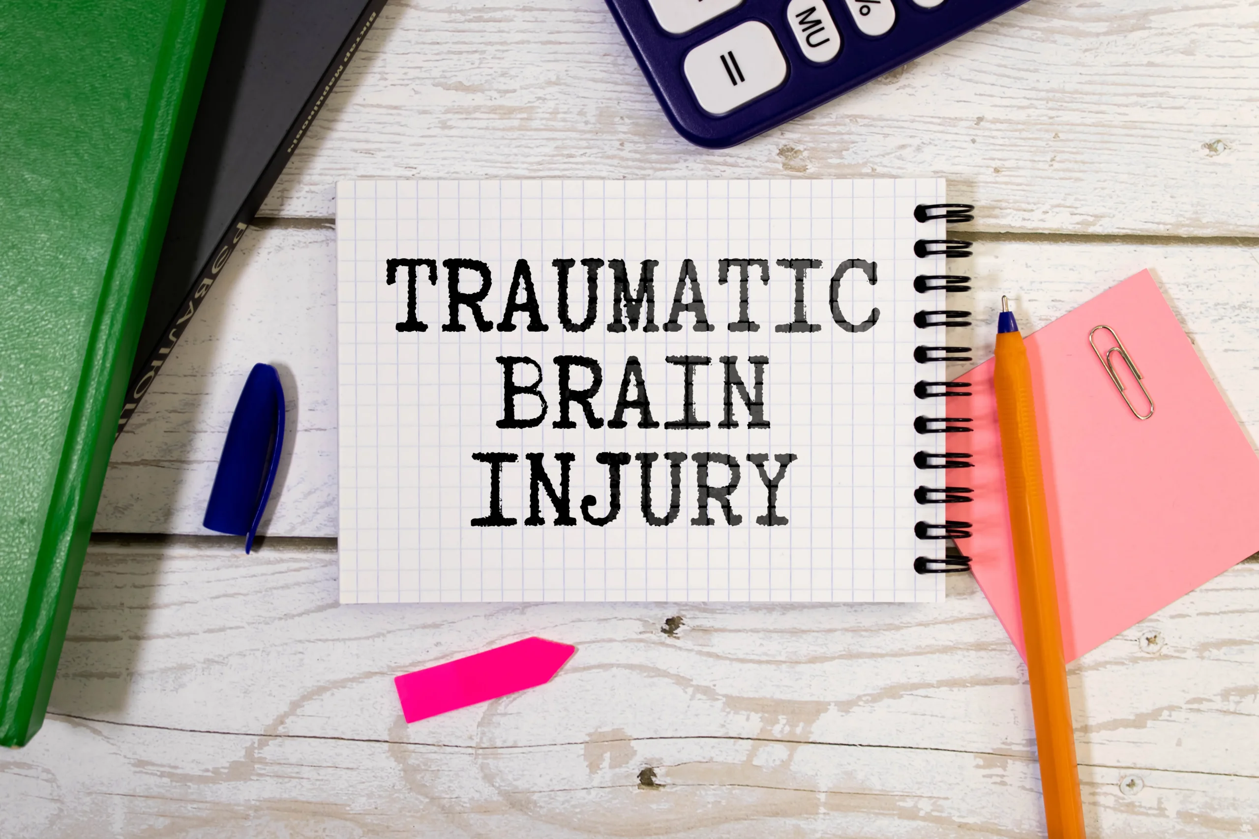 Notebook page with "TRAUMATIC BRAIN INJURY" text surrounded by office supplies on white wooden surface.