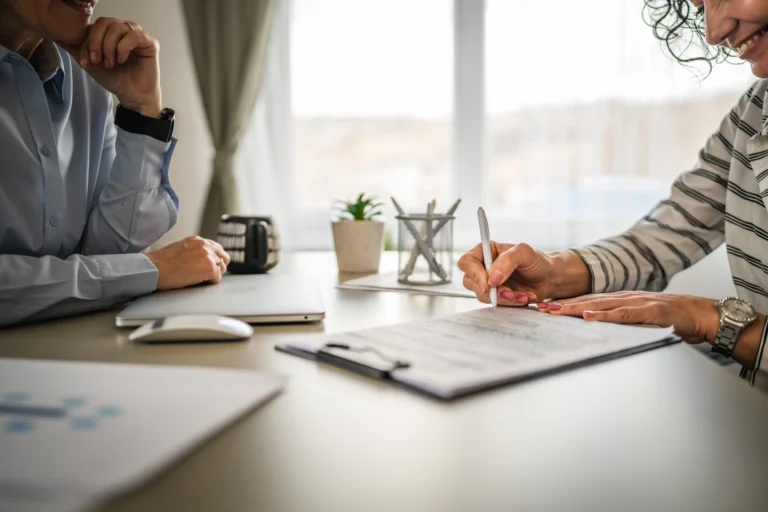 Two professionals at a desk with documents, a laptop, and office supplies near a window.